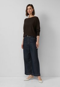 Femme debout portant un pull ample marron foncé, un jean bleu foncé à jambe large et des escarpins à talons bas blancs, devant un fond gris clair.