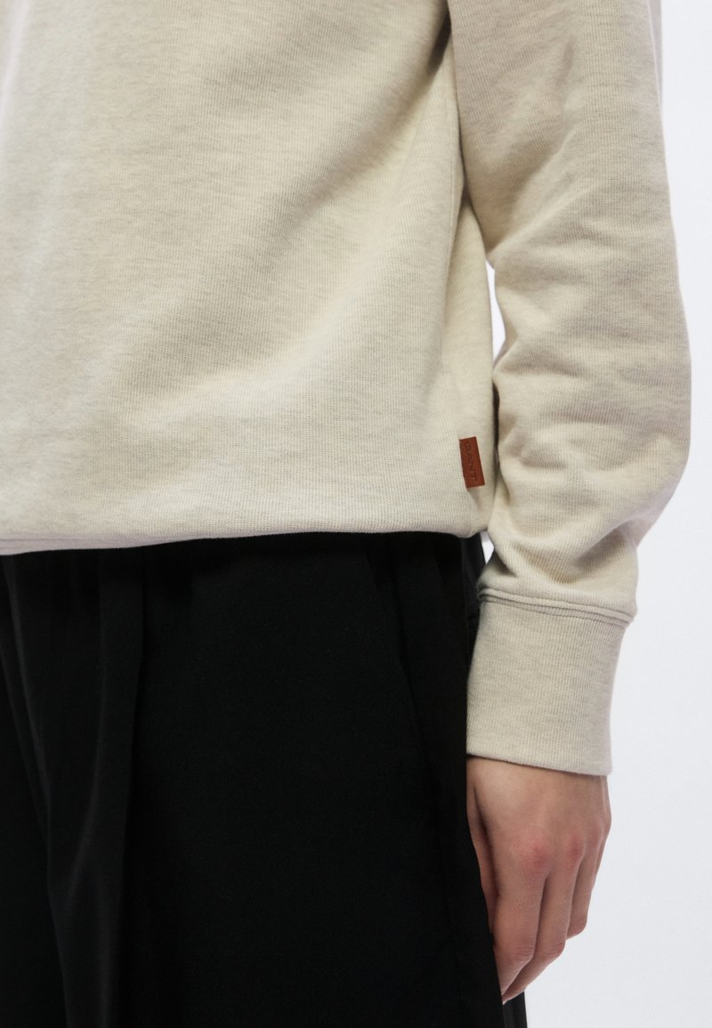 Light beige sweatshirt made of soft cotton, featuring long sleeves and a small brown label at the hem, paired with black pants.
