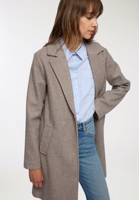 Donna con un cappotto di lana beige, una camicia a righe blu e bianche con bottoni e jeans azzurri, in piedi con un braccio piegato all'altezza della vita.