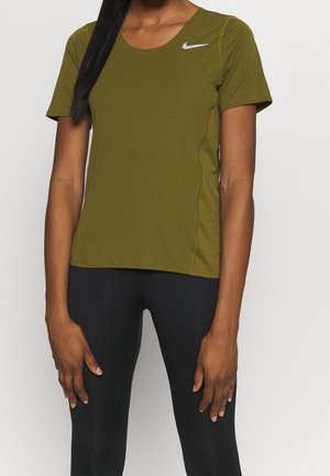 Vrouw draagt een olijfgroen Nike T-shirt met korte mouwen en zwarte legging, staat met ontspannen armen langs haar zij tegen een effen achtergrond.
