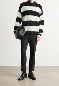 Sweter w paski czarno-kremowe z puszystego materiału, zestawiony z czarnymi spodniami i czarnymi sznurowanymi butami. Modelka trzyma okrągłą czarną torbę.