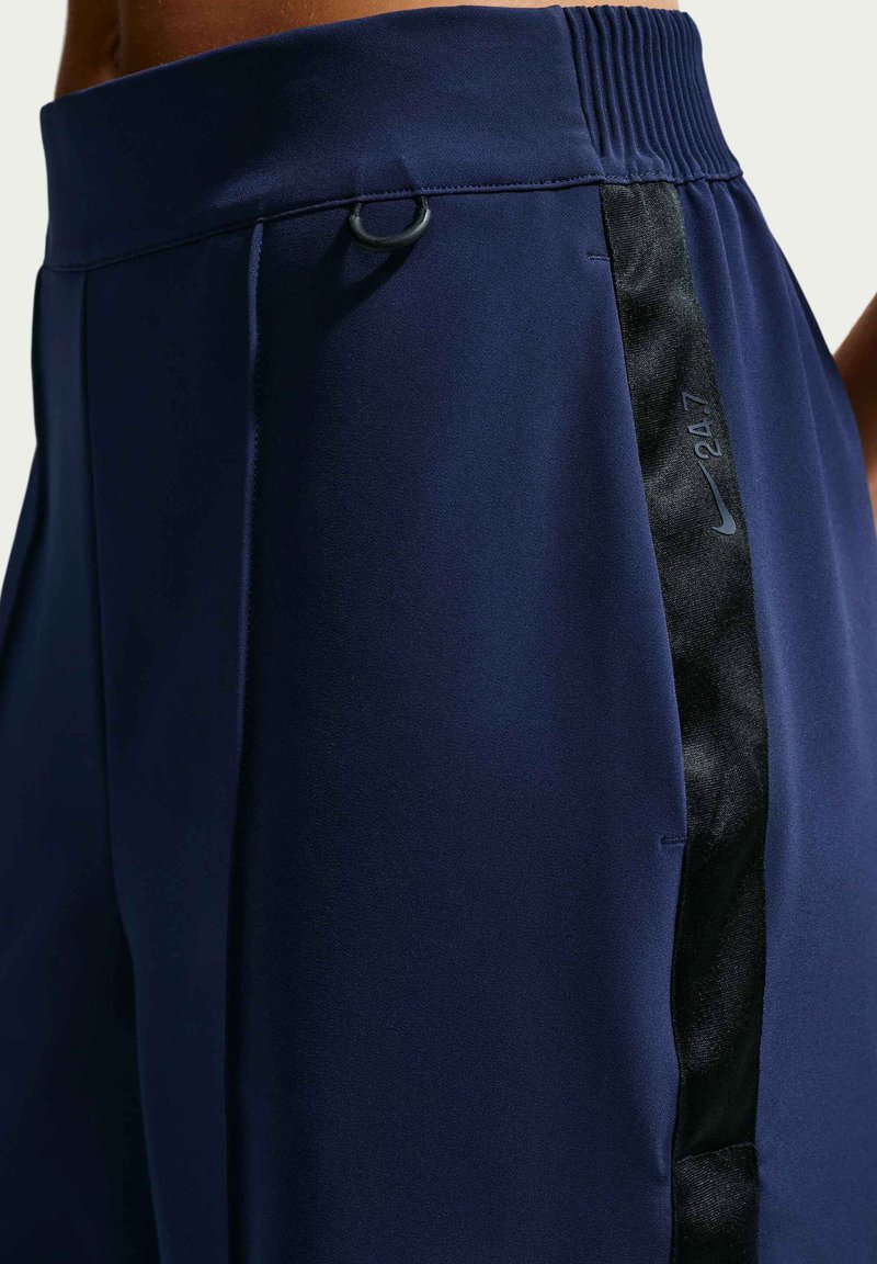 Nahaufnahme von marineblauen Sportshorts mit schwarzem Seitenstreifen, der kleinen reflektierenden "247"-Schriftzug und Nike-Swoosh-Logo nahe dem elastischen Bund zeigt.