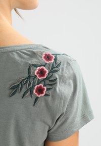 Grå kortærmet t-shirt med blomsterbroderi i pink og grøn på venstre skulder. Blødt stof med rund halsudskæring.