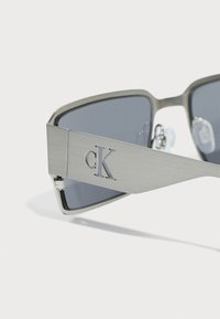 Ochelari de soare cu un cadru metalic gri, lentile întunecate gri și logo-ul gravat "cK" pe lateral. Design unghiular cu texturi netede.