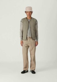 Person iført en beige bøttehat, olivengrøn quilted vest, grå langærmet bluse med ribbede detaljer, beige vide bukser og sorte sko med hvide striber.