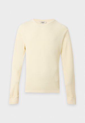 Homme portant une chemise beige clair à manches longues, un short bleu marine, des chaussettes blanches et des baskets marron, debout les mains dans les poches.