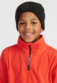 Orange fleece-polo med hög krage och svart dragkedja, ihop med en svart ribbad stickad mössa med en logotyp-patch på framsidan.