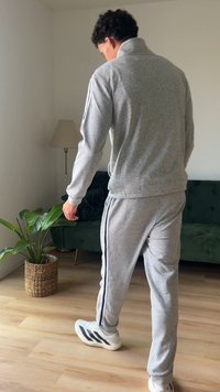Man in gray tracksuit and white sneakers walking on wooden floor near a green sofa, potted plant, and floor lamp in a bright room.