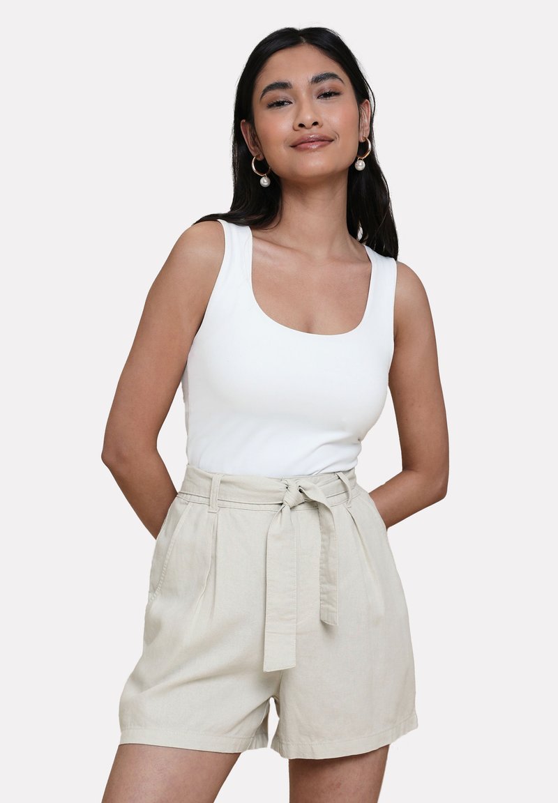 Top bianco senza maniche abbinato a pantaloni corti in lino beige con vita con cintura, tasche laterali e una vestibilità rilassata. Texture liscia, design casual.