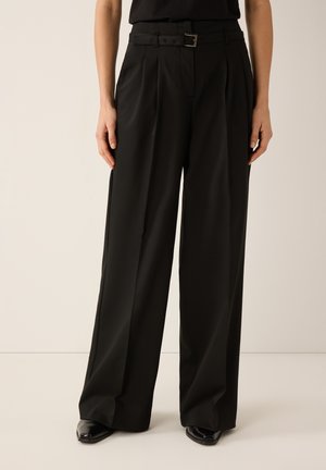 Frau trägt schwarze, hochtaillierte Hose mit weitem Bein und passendem Gürtel sowie schwarze spitze Schuhe, steht vor einem einfarbigen Hintergrund.