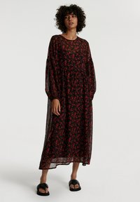 Schwarzes Blumenkleid mit roten Blumen, langen transparenten Ärmeln, gestuftem Design und rundem Halsausschnitt. Kombiniert mit schwarzen flachen Sandalen.
