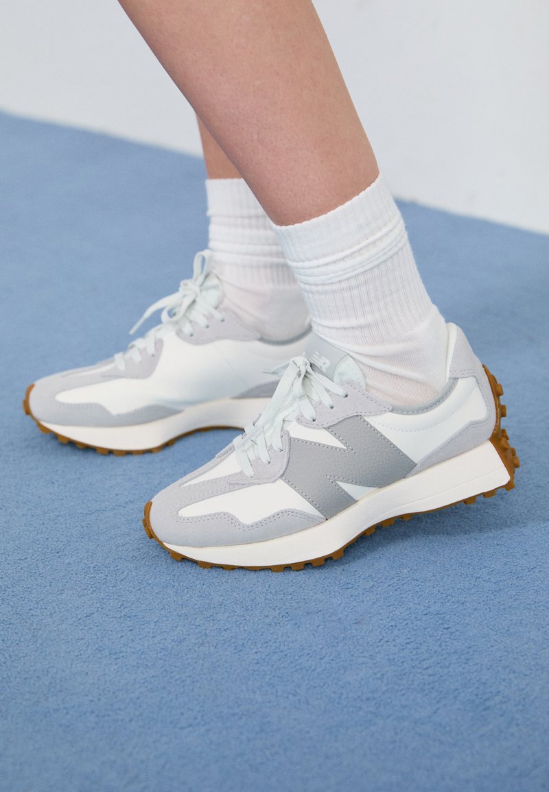 Scarpe da ginnastica grigio chiaro con accenti bianchi, tessuto texturizzato e suole in gomma, indossate con calzini bianchi a coste su una superficie blu.