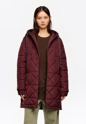 Kobieta ubrana w długą, pikowaną bordową kurtkę z kapturem, na koszulce brązowego koloru i jasnych zielonych spodniach, stojąca na tle jednolitego białego tła.