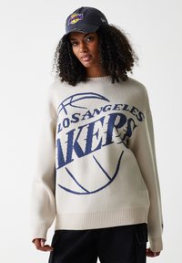 Pulover tricotat crem cu logo-ul albastru "LOS ANGELES LAKERS" și un design cu minge de baschet. Însoțit de o șapcă neagră cu logo galben și mov.