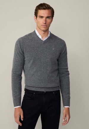 Jersey gris de mezcla de lana con escote en V, puños y dobladillo acanalados, combinado con una camisa blanca con cuello debajo del jersey, llevado con jeans oscuros.