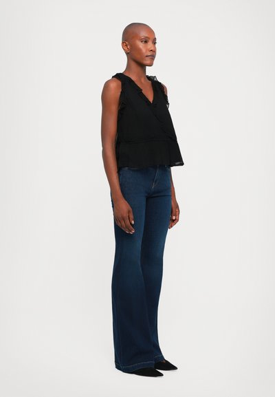 Zwarte mouwloze top met gerimpelde accenten, gelaagd ontwerp aan de zoom. Gecombineerd met uitlopende donkerblauwe jeans en puntige zwarte schoenen.