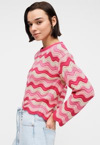 Νεαρή γυναίκα με κοντά σκούρα μαλλιά, που φοράει πουλόβερ πλεκτό με ροζ και κρεμ κυματιστό μοτίβο και ανοιχτό μπλε τζιν, με τα χέρια στις τσέπες.