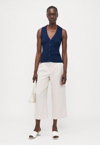 Donkerblauw geraffineerd gebreid mouwloos topje met een V-hals en een sluiting met knopen aan de voorkant, gecombineerd met roomkleurige wijde broek en witte slip-on sandalen.