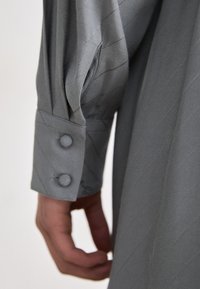 Manche en tissu gris avec un motif texturé diagonal, boutons décoratifs sur le poignet et une main partiellement visible tenant le tissu.