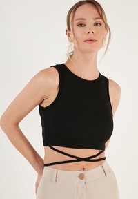 Must mustriba crop top kõrge kaelusega ja vööl ristikeerdega rihmad, koos heleda beežide pükstega, millel on puidust nupp.