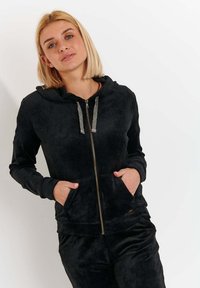 Felpa nera con zip realizzata in tessuto morbido, dotata di cappuccio, tasche frontali e lacci metallici. Texture liscia e vestibilità comoda.