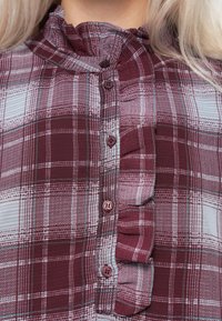 Lähedalt vaadatuna marooni ja helesinise ruudulise särk, millel on nööbid ja volditud äär nööpide ääres, kandes isik, kellel on blondid juuksed.