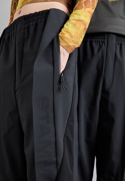 Czarne spodnie sportowe z oddychającego materiału, elastycznym pasem, boczną kieszenią na zamek błyskawiczny oraz perforowanym napisem "STAM" na jednej nogawce.