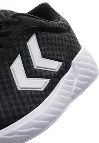 Svart sportskor med en texturerad textilovandel, vit gummisula och distinkta vita chevron-accenter på sidan.