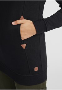 Svart sweatshirt med mjuk textur, med en triangulär läderdetalj på fickan och en liten logotypetikett vid fållen.