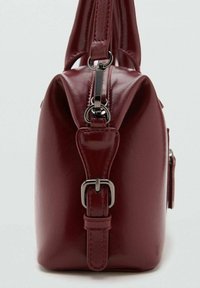 Pieni burgundinvärinen käsilaukku, jossa on sileä ja kiiltävä nahkapinta. Varustettu yläkantokahvalla, säädettävällä hihnalla ja hopeanvärisillä yksityiskohdilla.