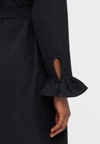 Μαύρο μανίκι φορέματος με ρυάκι στο τελείωμα και λεπτομέρεια κοπής. Υφή υφάσματος. Φαίνεται χέρι που κρατά το ύφασμα, αναδεικνύοντας το σχέδιο.