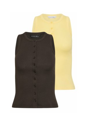 Donna in piedi con le mani dietro la schiena, indossa un top nero senza maniche con bottoni e jeans larghi azzurri su sfondo neutro.