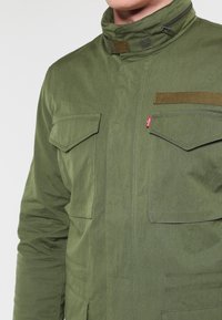 Grön militärjacka i slitstarkt material, med hög krage, fyra fickor med klaff och kontrasterande detaljer.