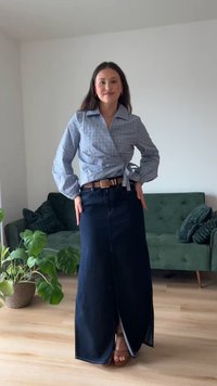 Giovane donna in piedi all'interno che indossa una blusa avvolgente blu con motivo, una lunga gonna scura con cintura e sandali marroni, con un divano verde e una pianta sullo sfondo.