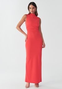 Rød ærmeløs maxikjole med høj hals, tætsiddende krop, rynket detalje i siden og glat tekstur, kombineret med lysefarvede hæle.