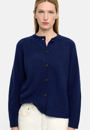 Kobieta w granatowym kardiganie na guziki z prążkowanymi mankietami i dołem, w połączeniu z kremowymi spodniami, stojąca na jednolitym tle.