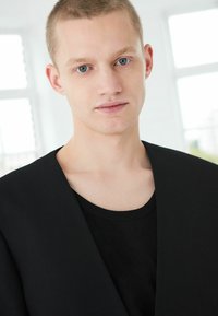 Jachetă neagră cu o suprafață texturată peste o cămașă neagră simplă. Modelul are părul scurt blond și ochii albaștri. Fundalul este luminos și neutru.