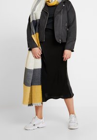 Abito midi nero, giacca in finta pelle grigia, sciarpa a fantasia check multicolore, sneakers bianche. Outfit casual a strati con diverse texture.