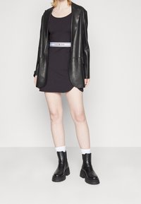 Musta nahkatakki sovitettu mustan mekon päällä, jossa on valkoinen logo-vyö. Mustat nilkkurit ja valkoiset sukat täydentävät asua.