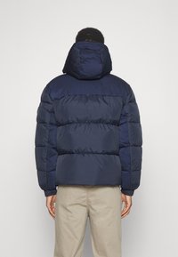 Marinblå pufferjacka med huva; har en quiltad design och elastiska ärmslut. Tillverkad av ett slätt, lätt material som ger värme och komfort.