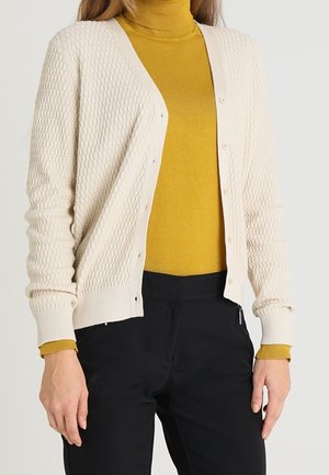 Un cardigan en tricot crème avec un motif texturé, doté d'un décolleté en V profond, de fermetures à boutons sur le devant et de poignets côtelés, superposé à un col roulé moutarde.