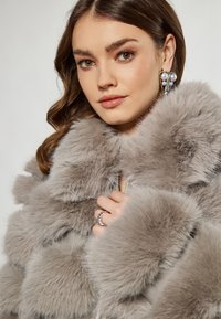 Šedý kabát z umělé kožešiny s nadýchanou texturou; má vysoký límec a zipové zapínání. Modelka ho drží zavřený prstenem na jedné ruce.