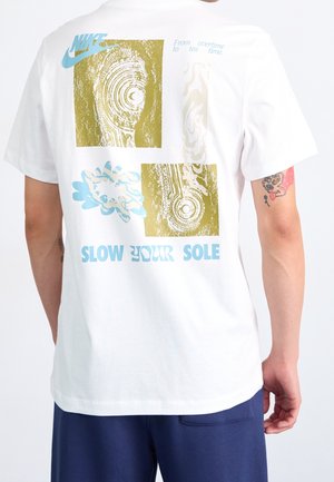 Hvid T-shirt med beige og blå abstrakte tryk, Nike-logo, tekst "Slow Your Sole,", båret af en person med en tatovering på armen.