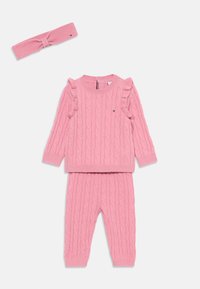 Completo maglione e pantaloni rosa lavorati a maglia con motivo a treccia, dettagli arricciati sulle maniche del maglione e fascia abbinata. Texture morbida.