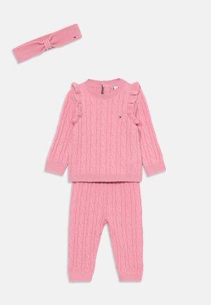 Ensemble de pull et pantalon en laine rose avec un motif en câble, détails froncés sur les manches du pull et bandeau assorti. Texture douce.