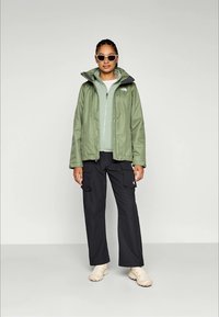 Jachetă impermeabilă de culoare verde deschis, cu căptușeală gri, cu fermoar și glugă, purtată peste un top verde deschis și pantaloni cargo negri; pantofi albi.