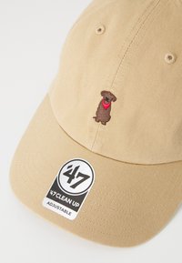 Gorra de algodón beige con correa ajustable. Presenta una silueta de perro marrón bordada en la parte frontal con un pañuelo rojo. Etiqueta negra mostrada.