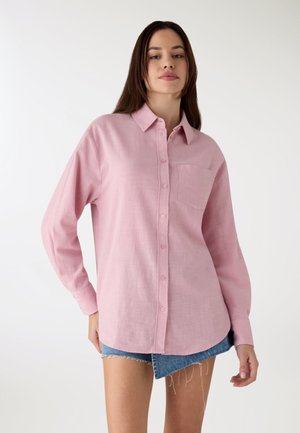Camicia rosa chiaro a manica lunga con bottoni e tasca; tessuto testurizzato; vestibilità comoda; abbinata a pantaloncini di jeans con bordi sfrangiati.