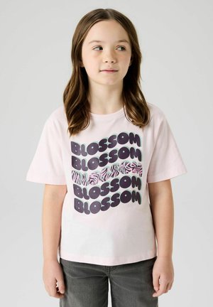 Ung pige med brunt hår iført en lyserød T-shirt med fed tekst "BLOSSOM" og zebrastribemønster, kombineret med mørke bukser.