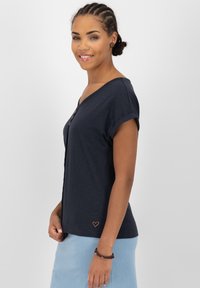 Camicia a maniche corte blu navy con scollatura a V, dettaglio dei bottoni in legno e un piccolo logo a forma di cuore sull'orlo. Tessuto morbido con una vestibilità comoda.